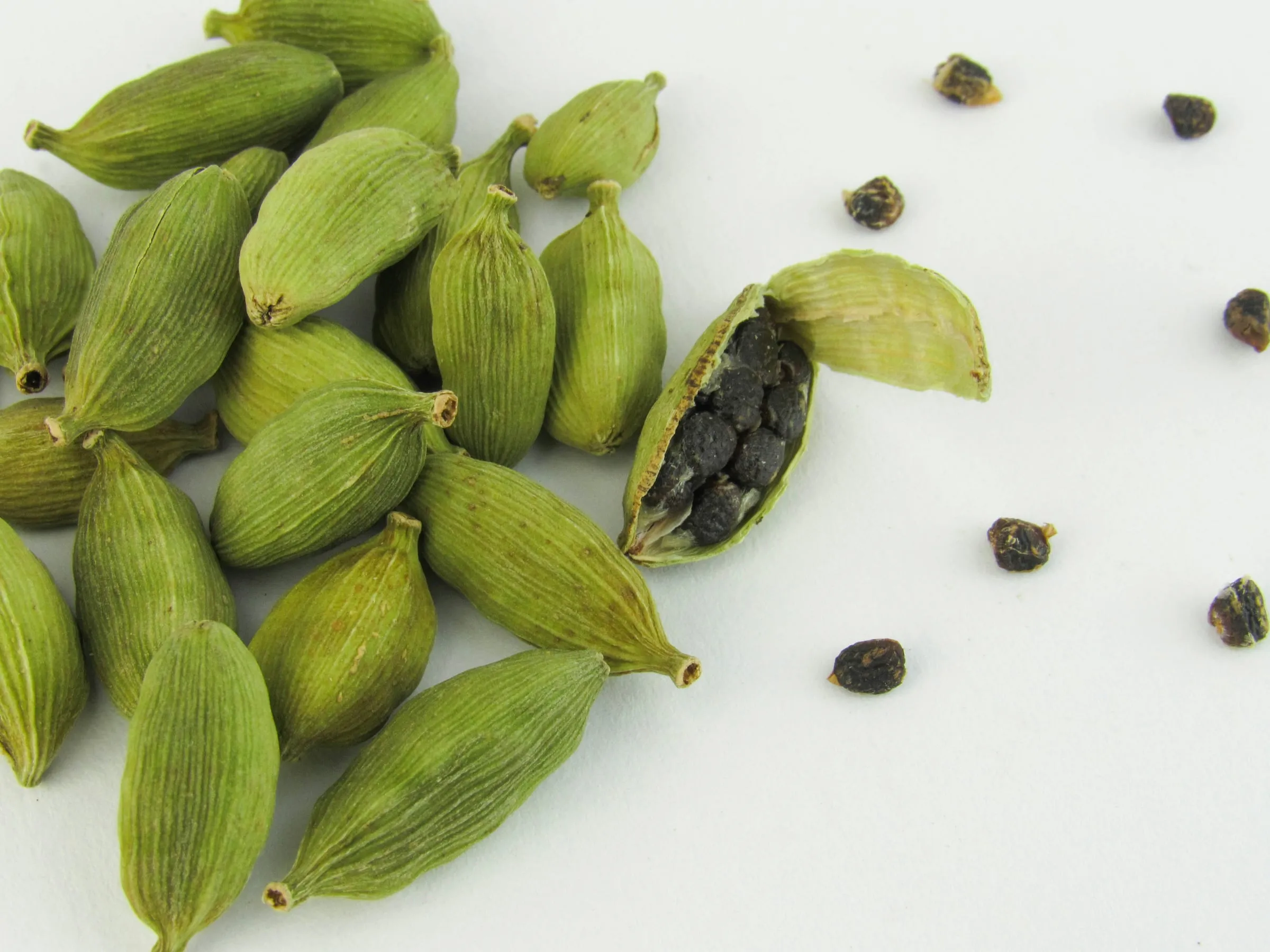 Green cardamom capsules on a white surface, one split open to show the dark seeds inside, with loose seeds scattered beside it