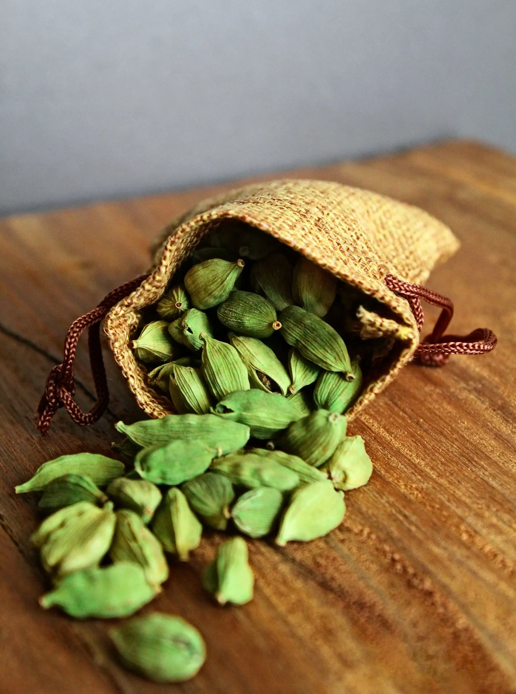 A small drawstring jute pouch tipped on its side on a wooden board, cardamom capsules spilling out of it