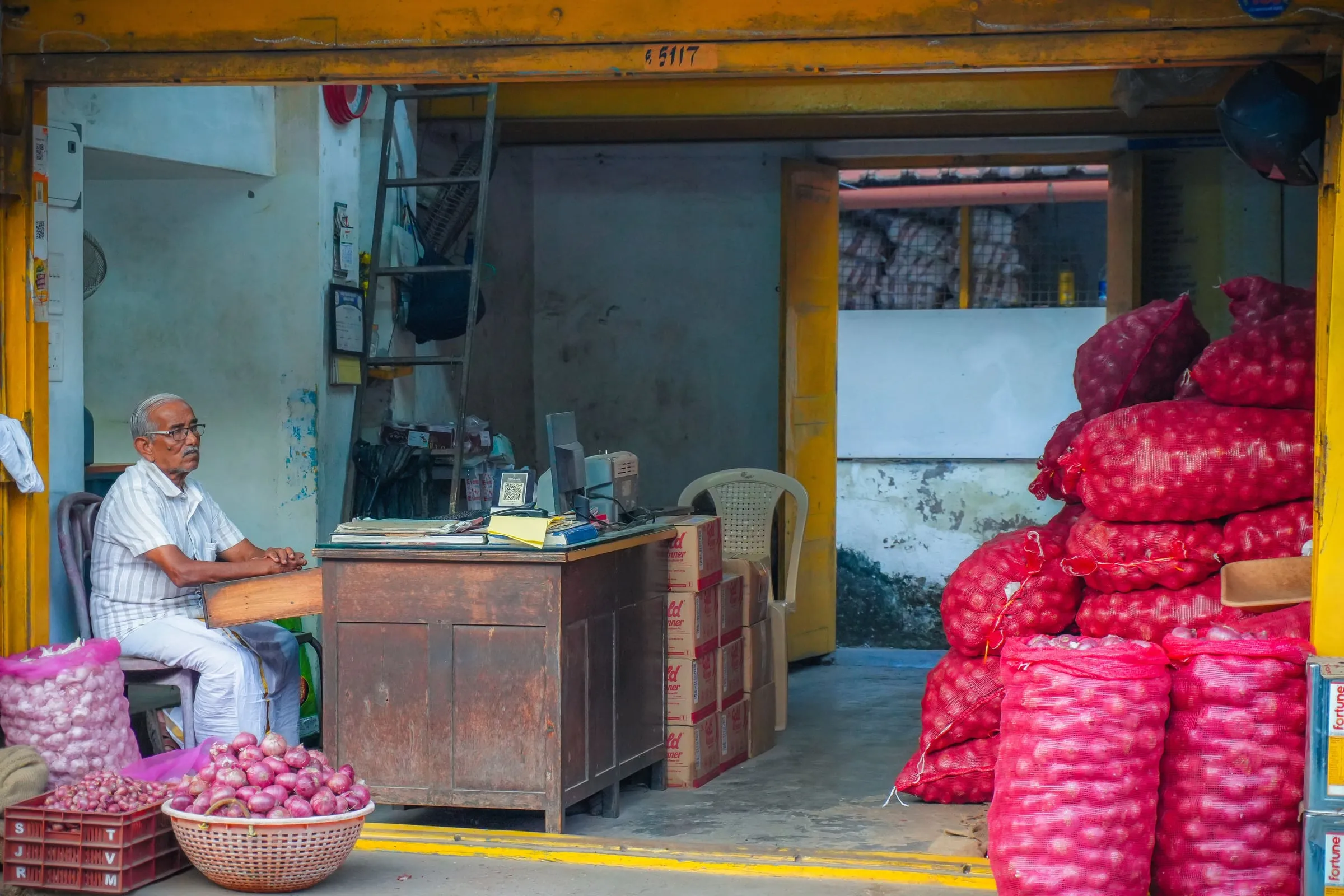 A wholesale onion shop with a trader sitting at a desk, mesh sacks of red onions stacked to one side and a basket of loose onions at the front