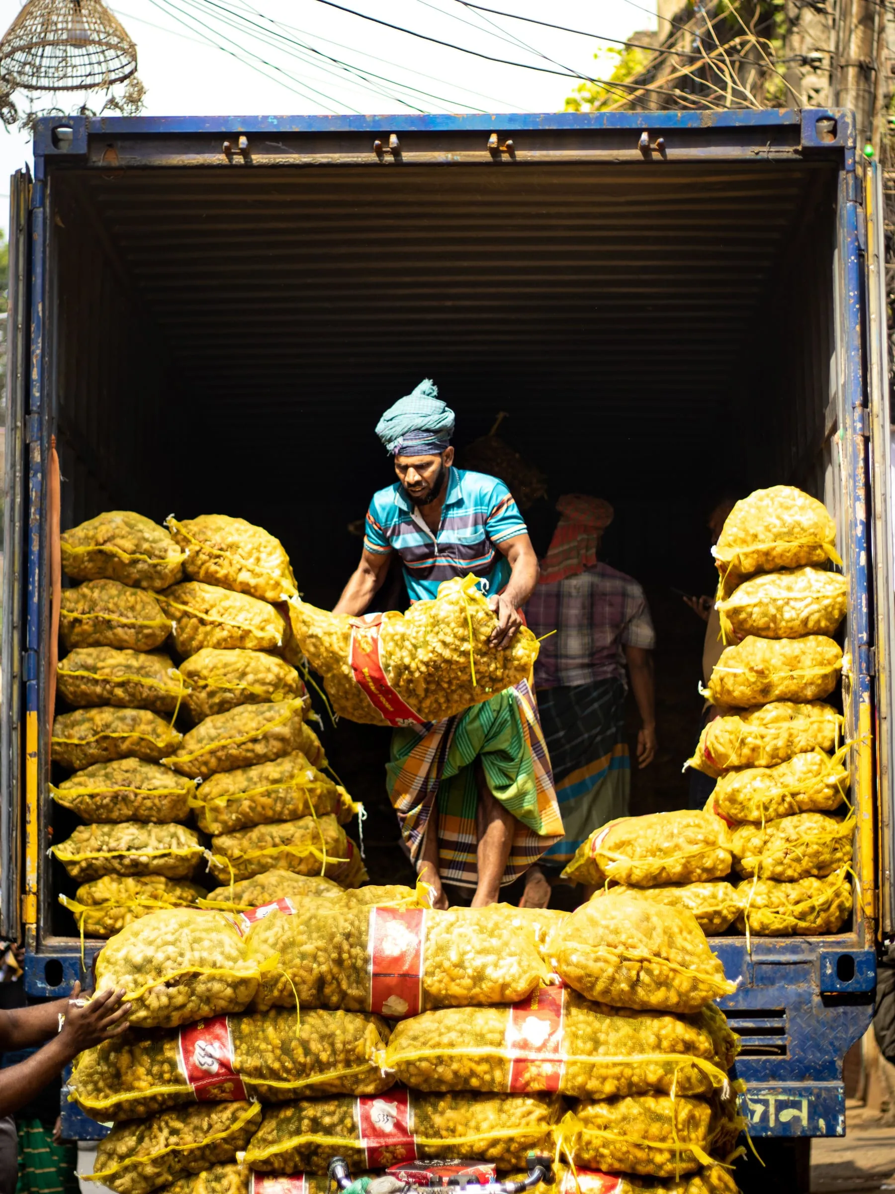 A worker lifting a mesh sack of ginger inside a shipping container, sacks stacked on both sides of him