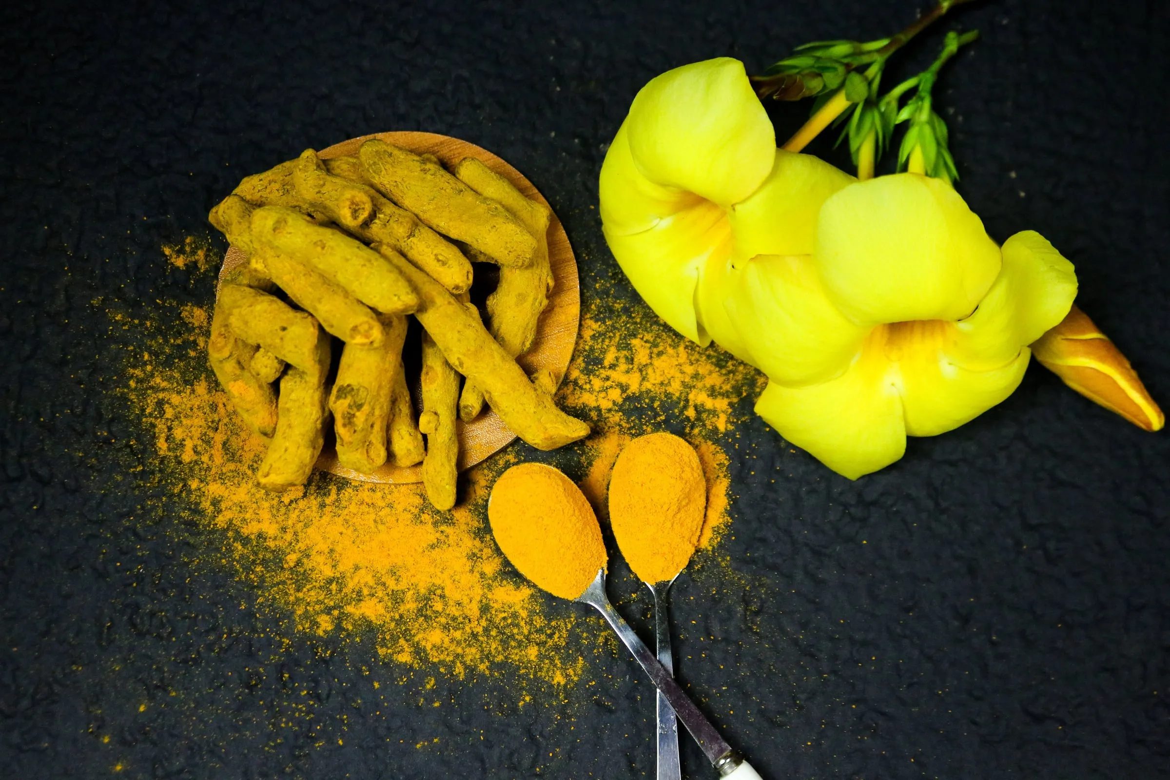 Dried turmeric fingers heaped on a round wooden dish beside two spoons of ground turmeric, powder scattered across a dark stone surface, with yellow flowers to one side